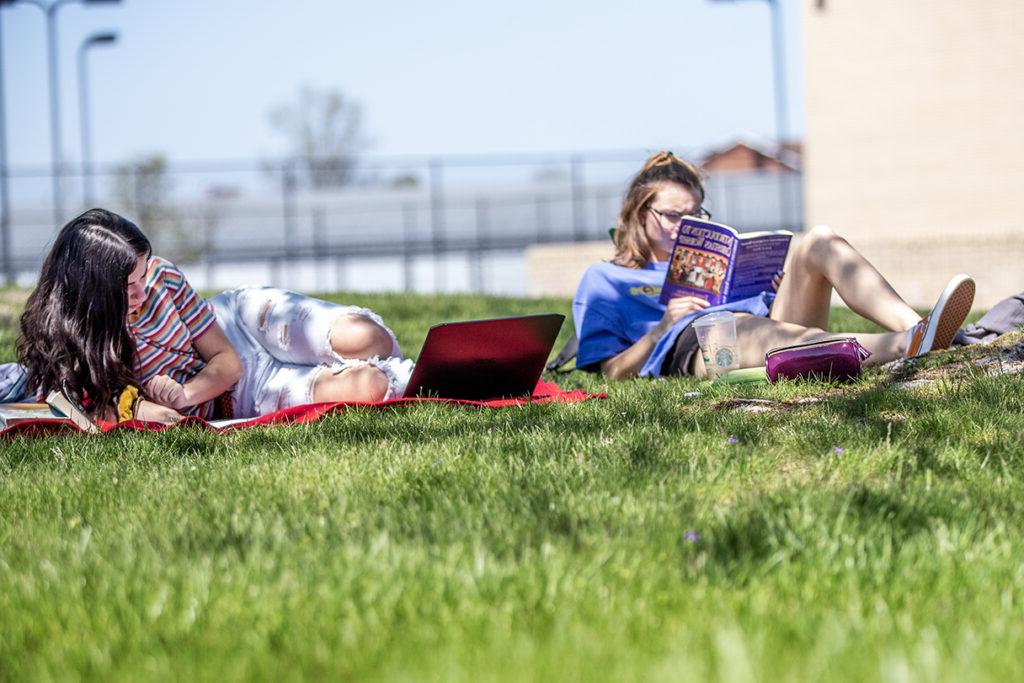 Two people lying on the grass; one is reading a book 和 the other is using a laptop.